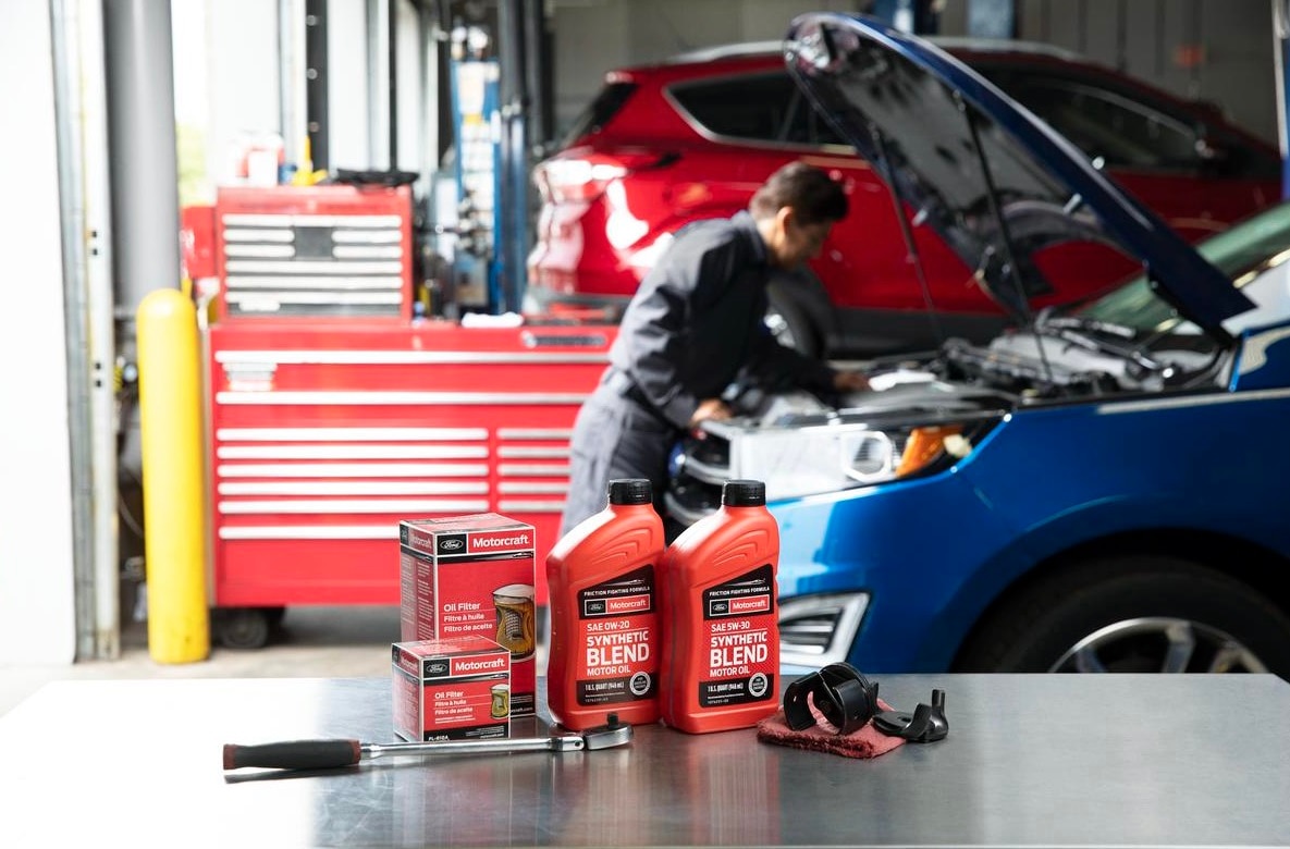 Motocraft brand products sit on a table with a shop technician working under a vehicle's hood in the background.